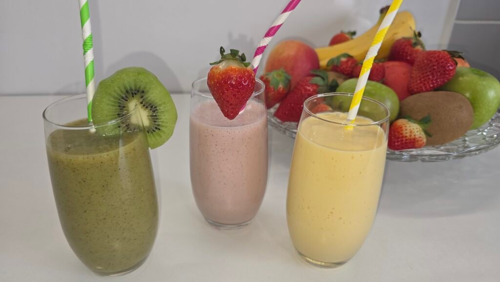 Trois verres de smoothies colorés (rouge, jaune, vert) sur une table de cuisine, avec des fruits frais à côté, illustrant des recettes de smoothies healthy faciles par Anne Maman Gâteaux.