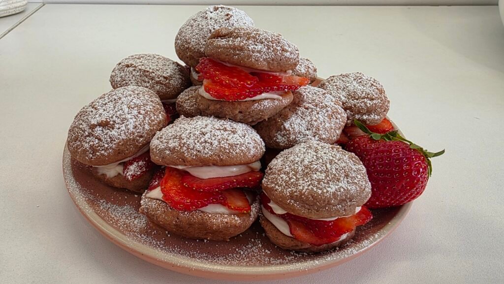 Deux cookies Whoopies à la fraise saupoudrés de sucre glace, garnis de crème blanche et morceaux de fraises, posés sur une belle assiette de présentation rose.