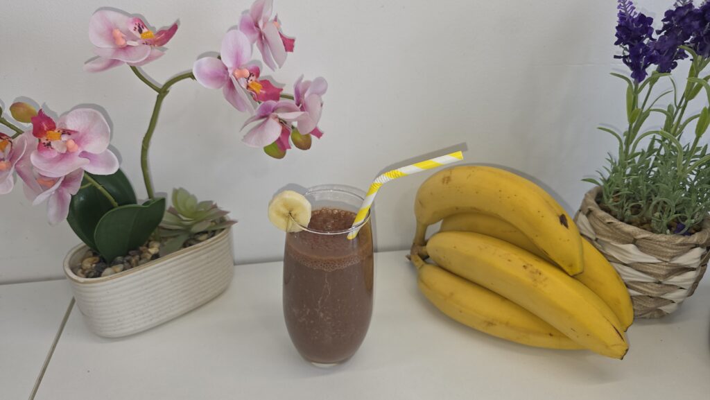 Un grand verre de smoothie banane chocolat avec des bananes à droite, des carrés de chocolat et une orchidée décorative à gauche.