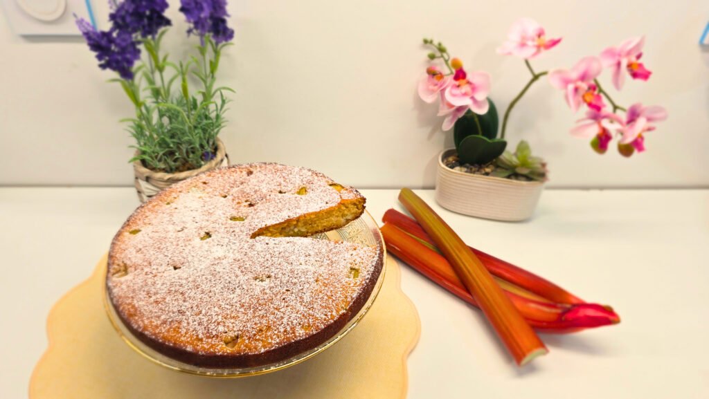 Gâteau moelleux à la rhubarbe sur un plat de service en verre avec des branches bien rouge de rhubarbe sur la droite et deux élément de décor lavande et orchidées en arrière plan.