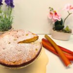Gâteau moelleux à la rhubarbe sur un plat de service en verre avec des branches bien rouge de rhubarbe sur la droite et deux élément de décor lavande et orchidées en arrière plan.