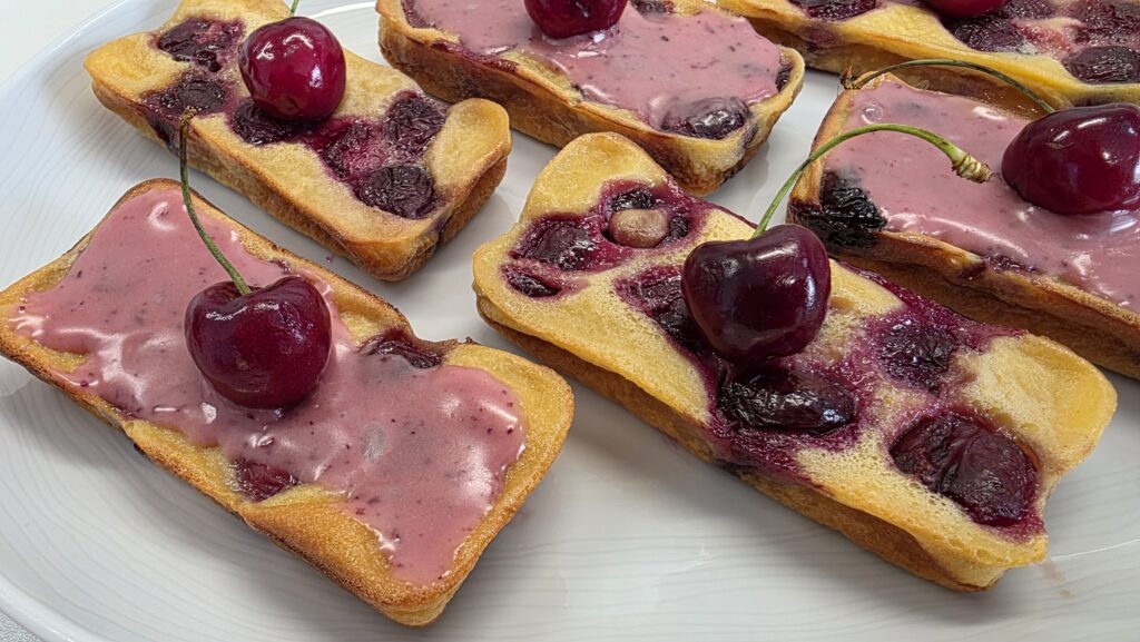 Clafoutis individuels en gros plan de forme rectangulaire (mini cake) avec pour l'un un glaçage à la cerise, surplombés d'une jolie cerise fraîche.