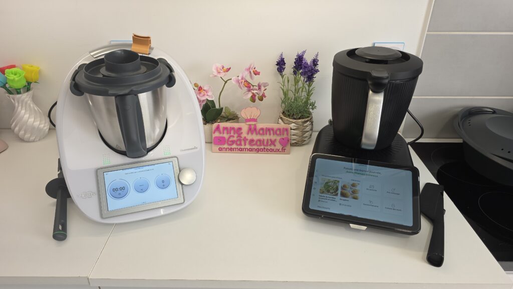 Thermomix Tm6 à gauche sur un plan de travail et Thermomix Tm7 à droite, au centre le logo : Anne Maman Gâteaux avec une orchidée et une lavande décoratives.