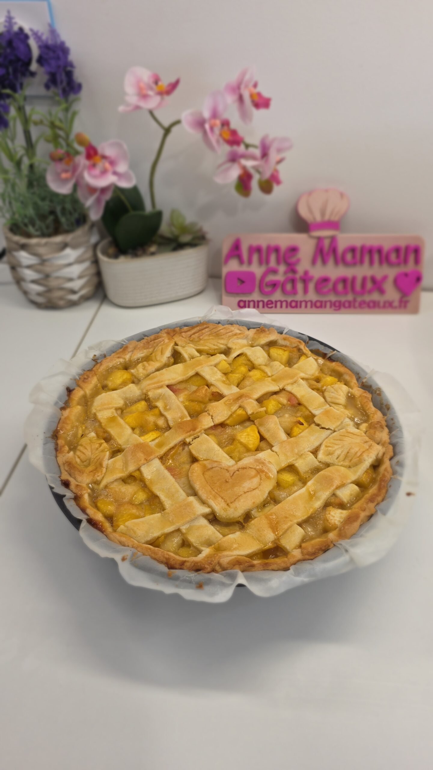 Tarte aux pêche sur un plan de travail avec un decor de lavande, orchidées et logo Anne maman gâteaux en fond.