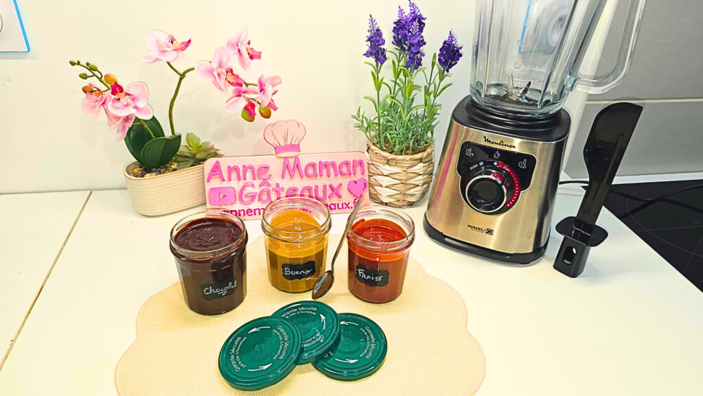 3 pots de pâtes à tartiner maison : chocolat/noisettes, façon Bueno et à la fraise. sur un plan de travail avec un blender Moulinex Perfectmix à droite et le logo Anne Maman Gâteaux en fond