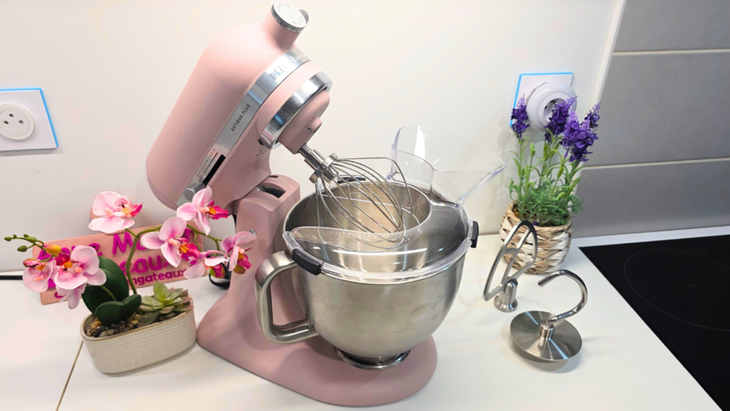 robot pâtissier KitchenAid Artisan Plus ouvert sur un plan de travail blanc avec ses accessoires : le batteur plat et le crochet pétrisseur.
