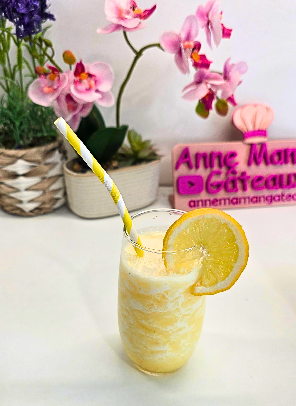 un verre haut rempli d'un smoothie jaune vif à l'ananas/coco avec une paille jaune et une rondelle de citron accrochée au bord du verre