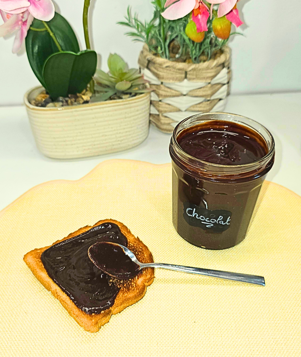 une tranche de pain de mie grillée recouverte de pâte à tartiner maison au chocolat avec encore la cuillère posée dessus, un pot de pâte à tartiner maison plein ouvert, le tout posés sur un set de table jaune pâle et en fond deux plantes artificielles orchidée et lavande.