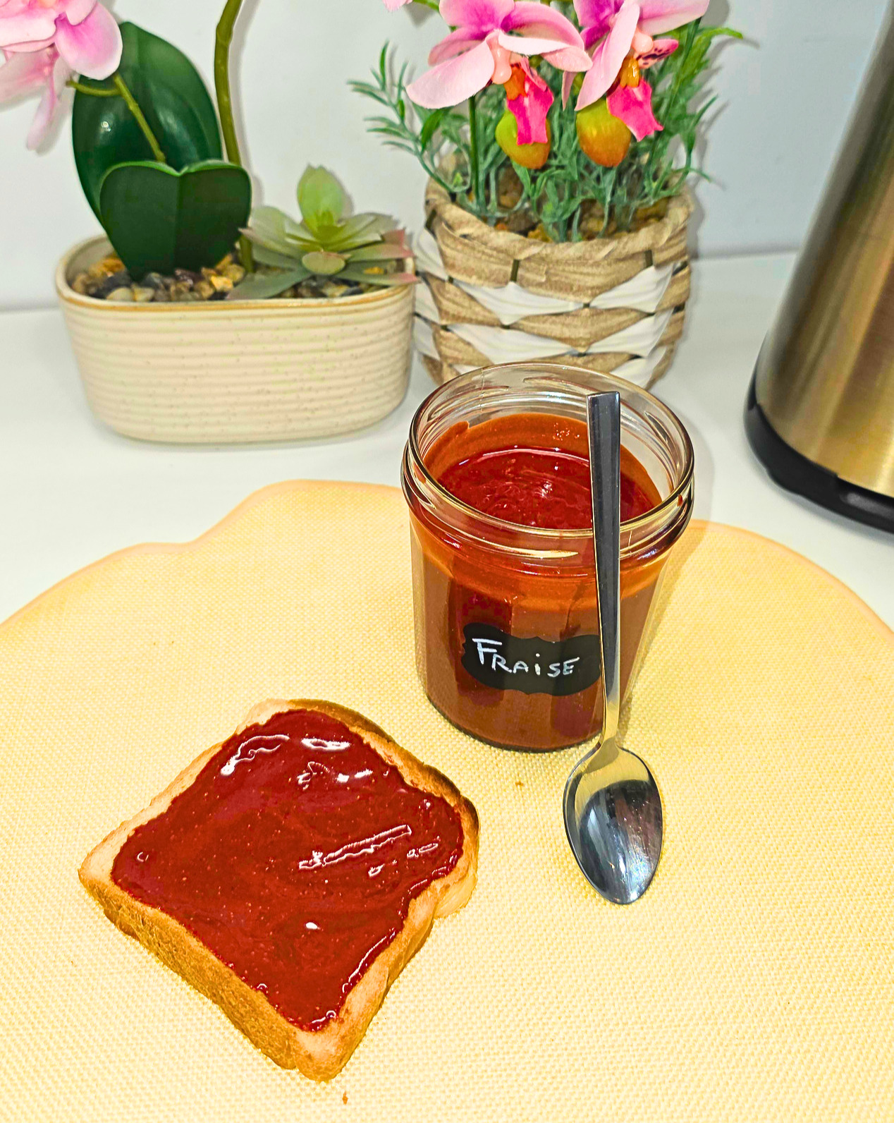 en gros plan : une tranche de pain de mie grillée tartinée de pâte à tartiner à la fraise, avec une cuillère en inox brillante et un pot de pâte à tartiner à la fraise posé sur un set de table jaune pale sur un plan de travail de cuisine en fond : deux pots de plantes artificielles orchidée et lavande.