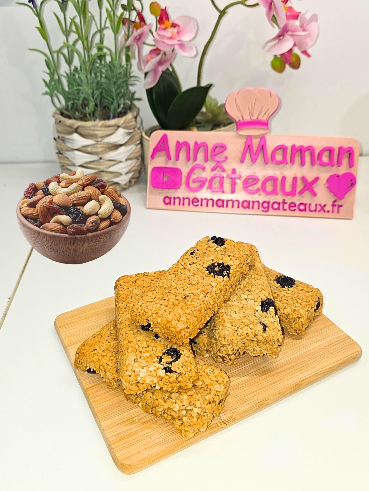 6 barres de céréales empilées sur une petite planche à découper en bois avec en fond un bol d'oléagineux averc raisins secs, logo Anne Maman Gâteaux